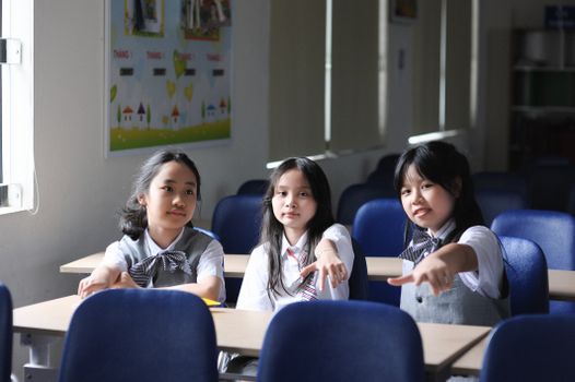 Three young students in a classroom pointing happily at the camera. Bright learning environment.