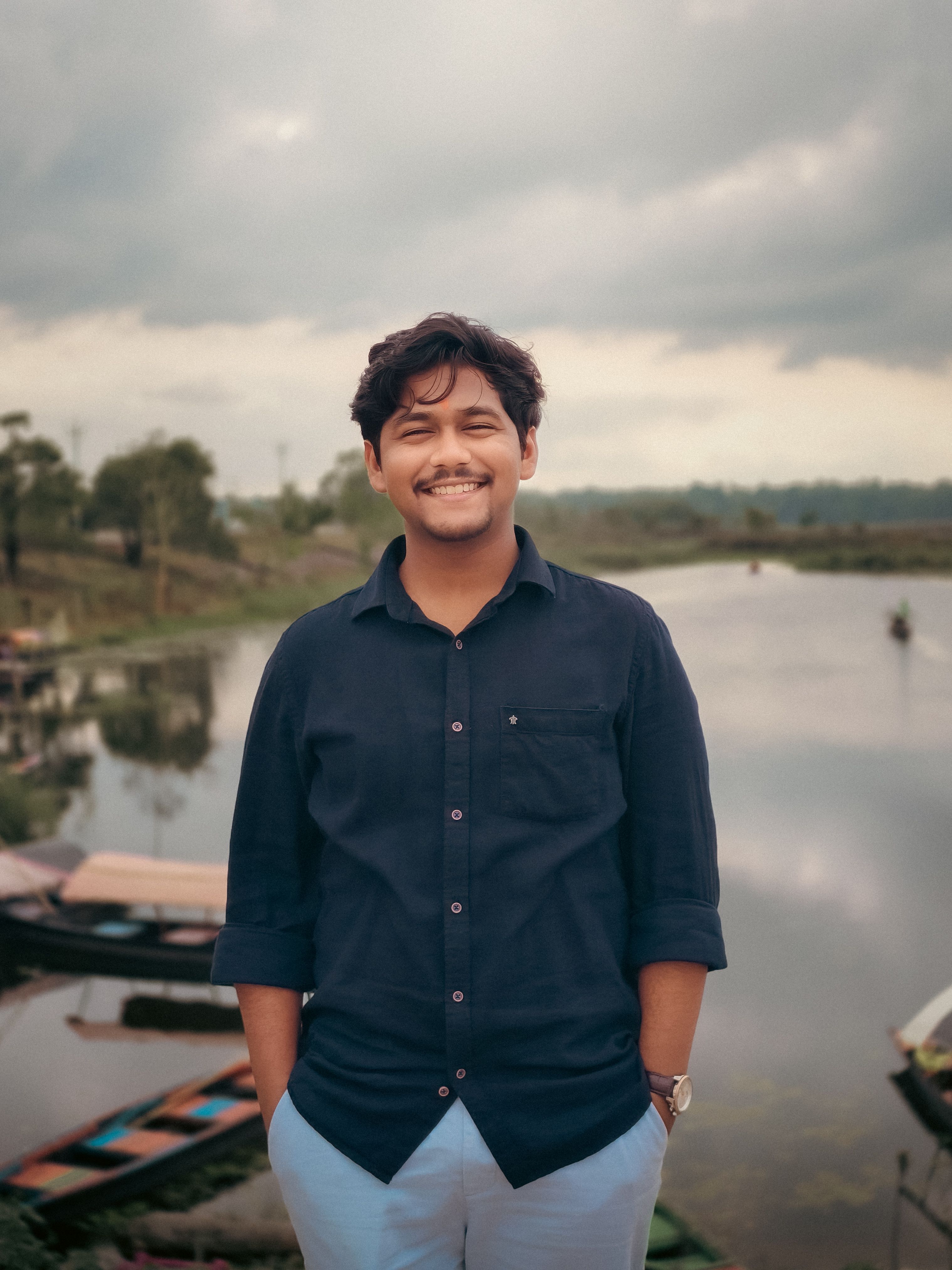 A cheerful young man poses by a serene lake under a cloudy sky.