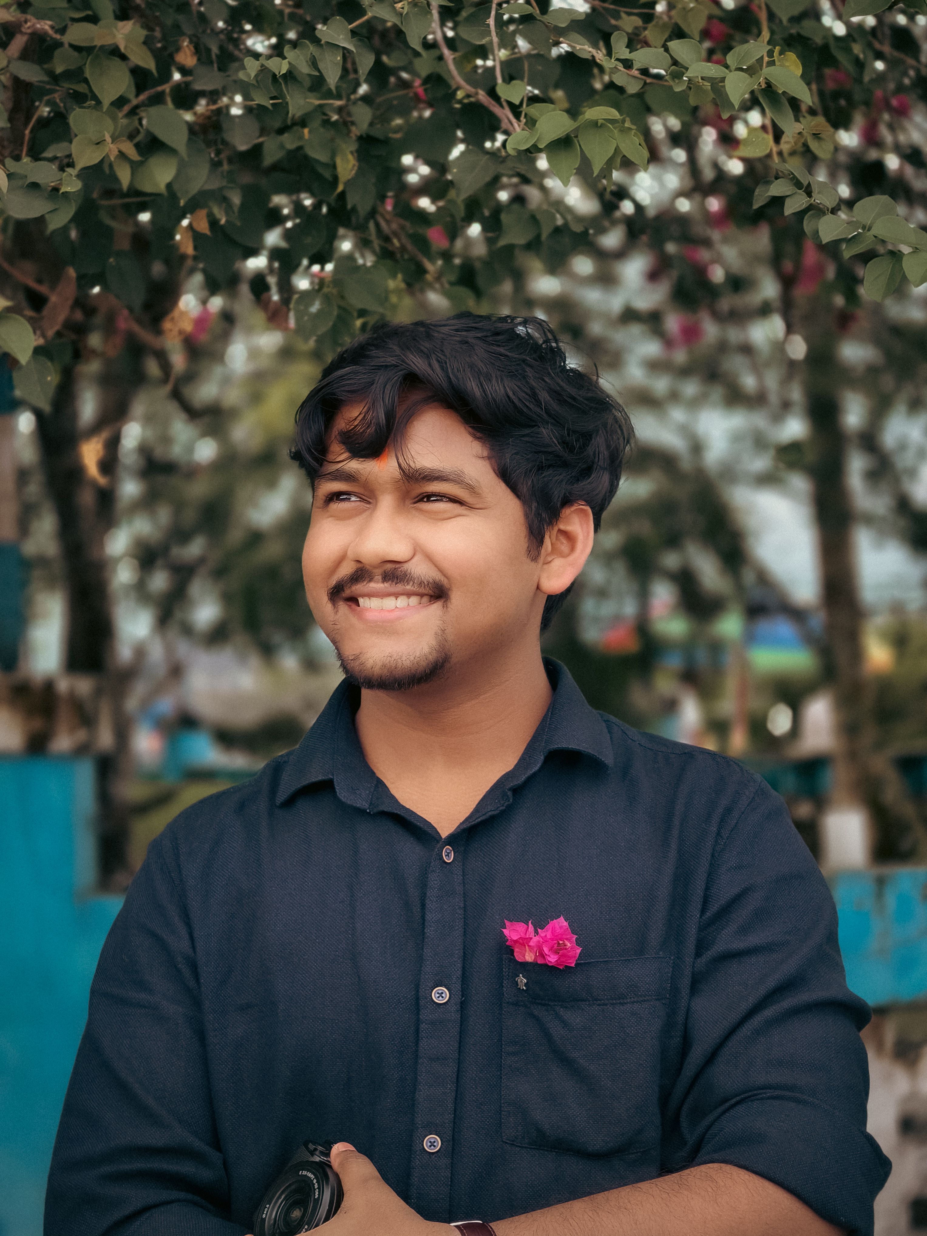 A young man smiling outdoors with a pink flower pinned on his shirt.