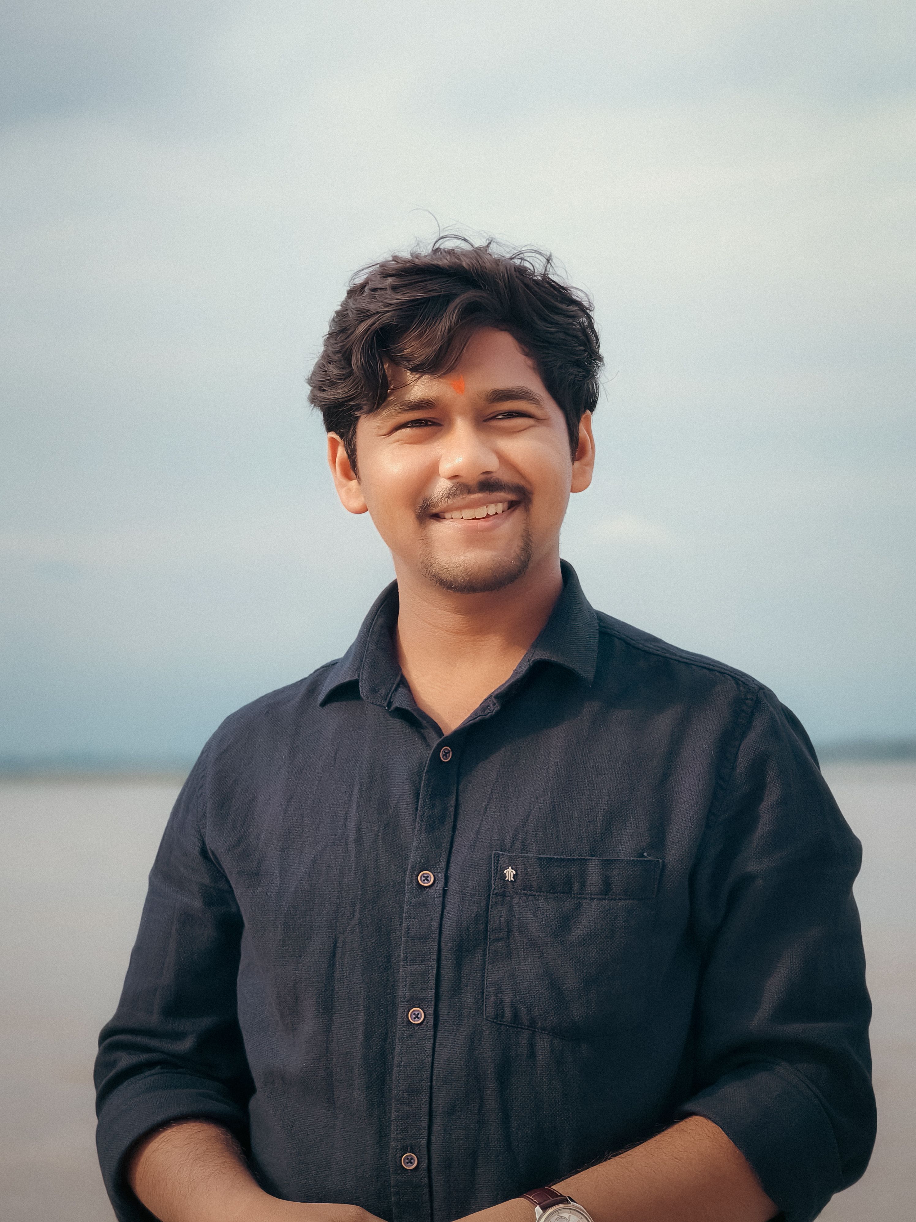 A smiling man stands outdoors with a scenic background of water and sky, conveying a warm and relaxed atmosphere.
