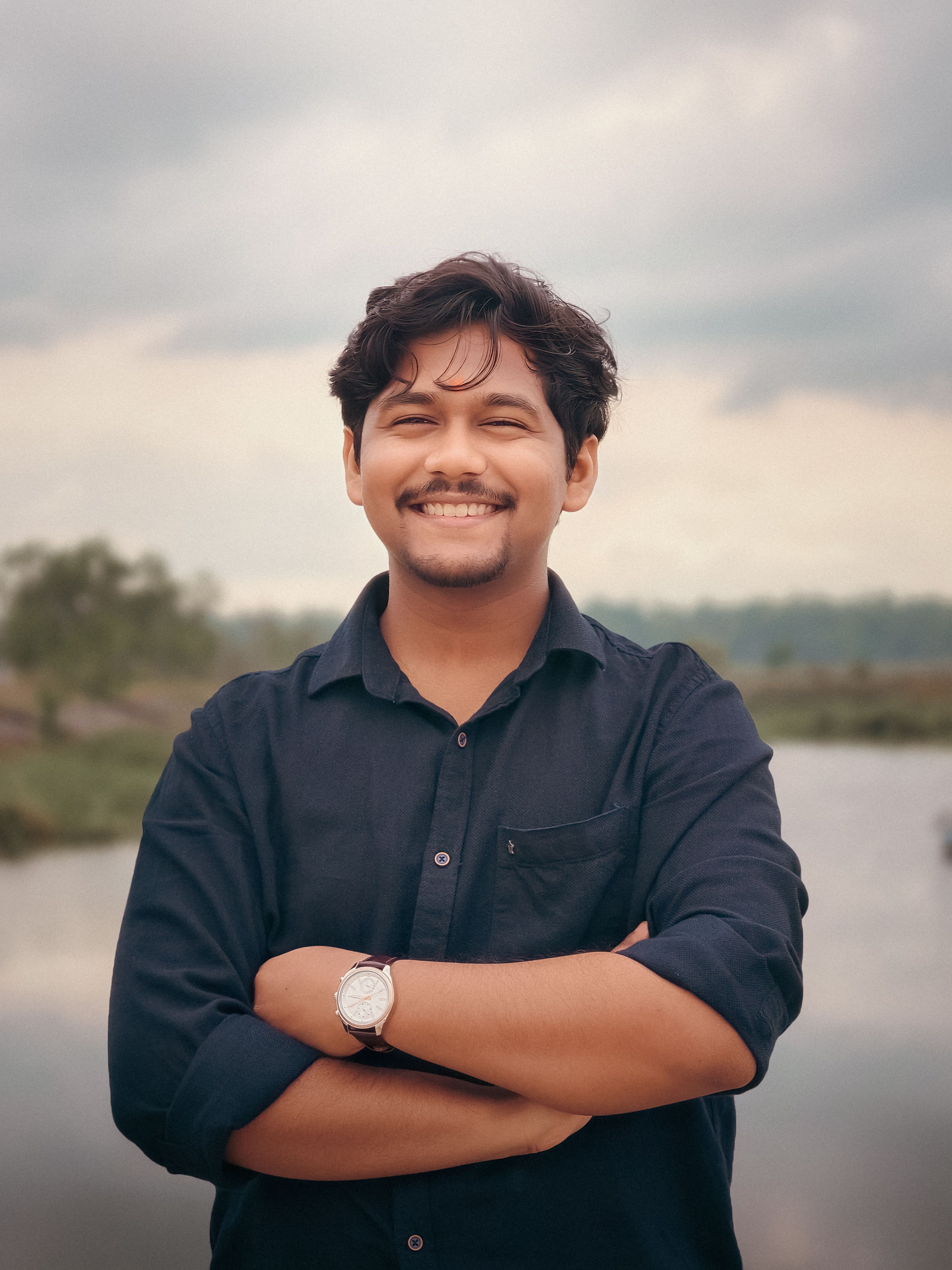 Smiling young man in casual attire with arms crossed standing by a scenic river under a cloudy sky.