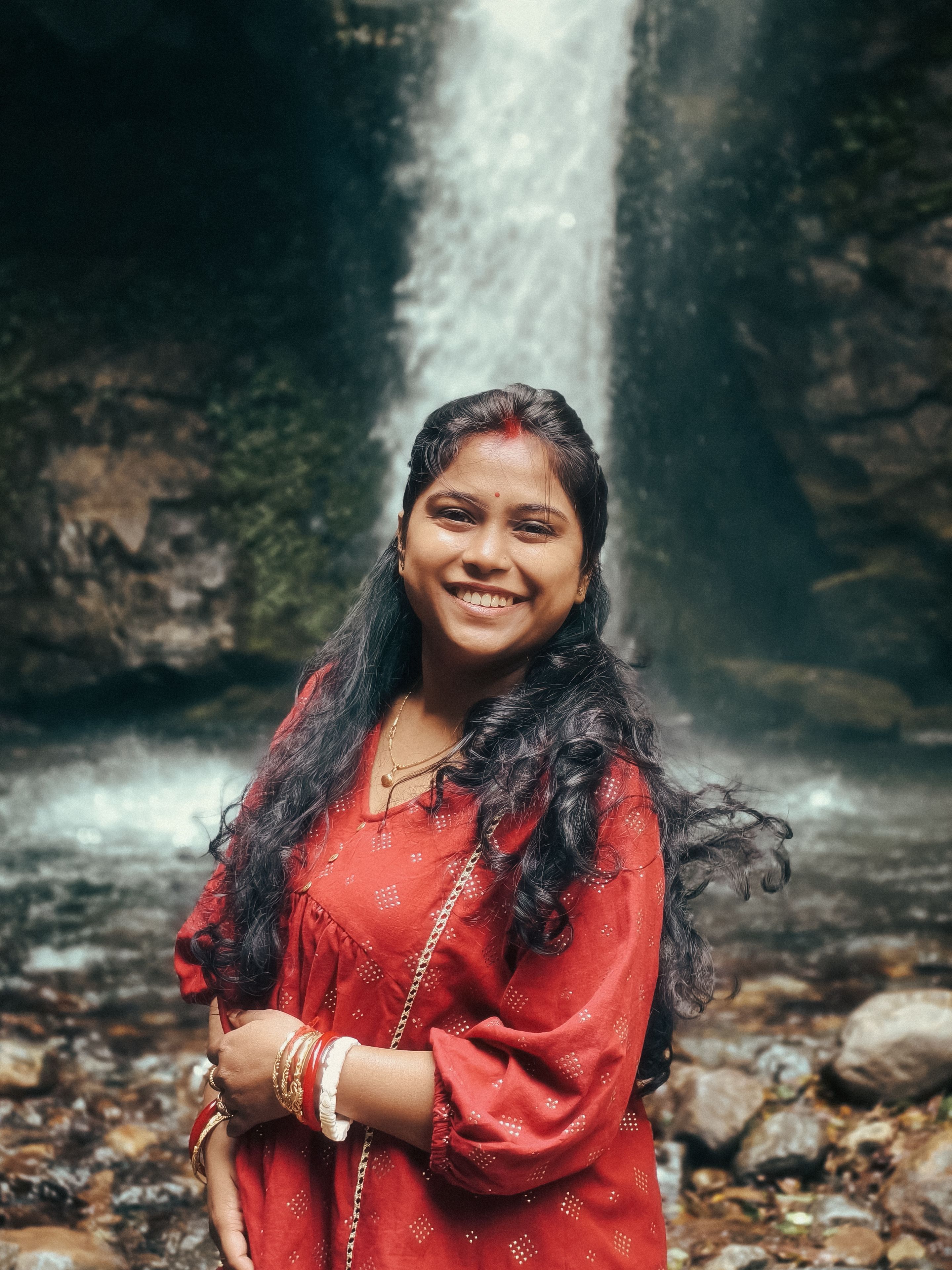Joyful woman in red traditional attire standing by a flowing waterfall
