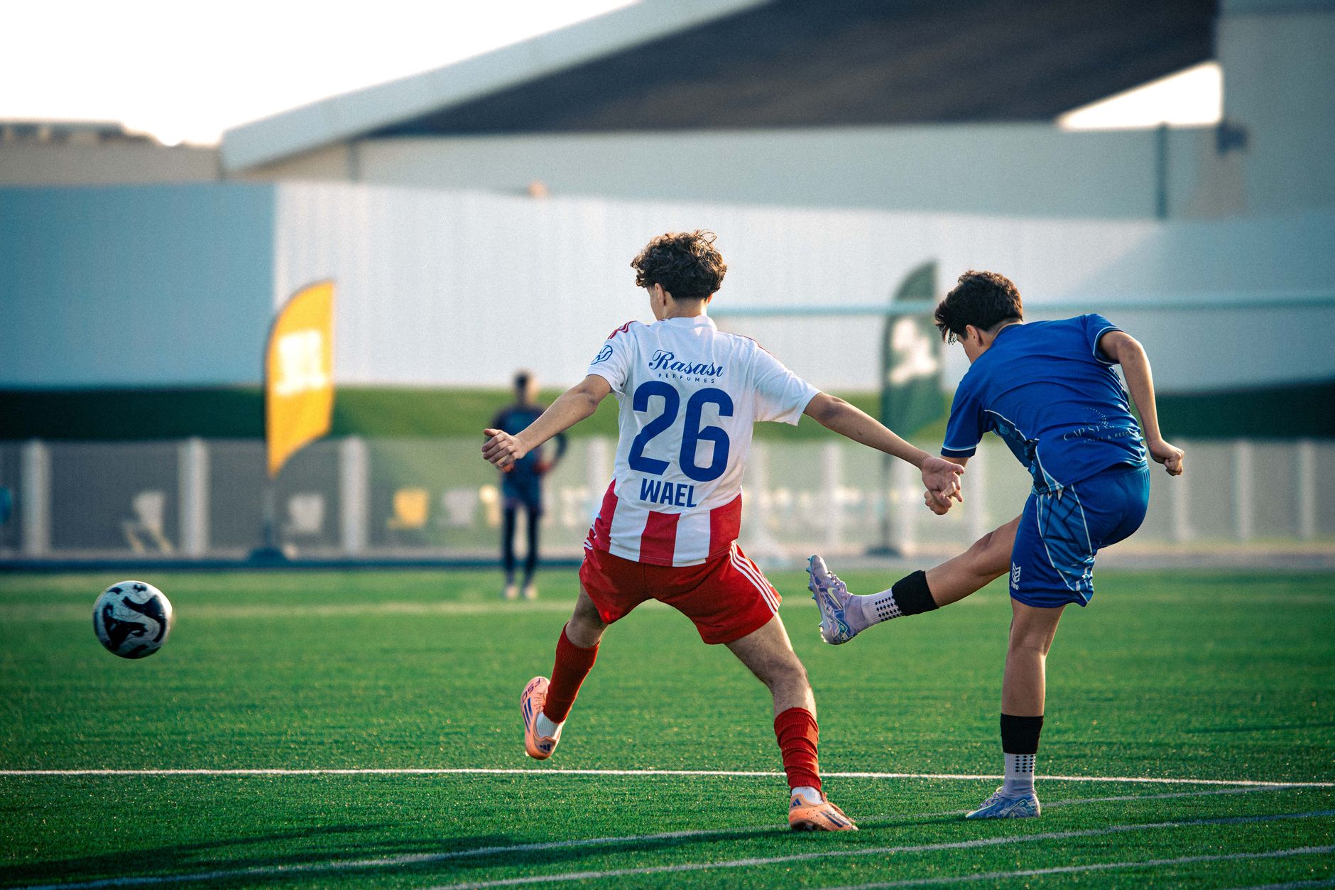 Two teenage boys playing soccer on a green field, mid-action kick under bright daylight.