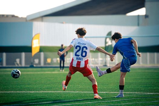 Two teenage boys playing soccer on a green field, mid-action kick under bright daylight.