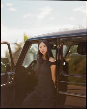 Elegant young woman in black outfit next to a luxury black car outdoors.