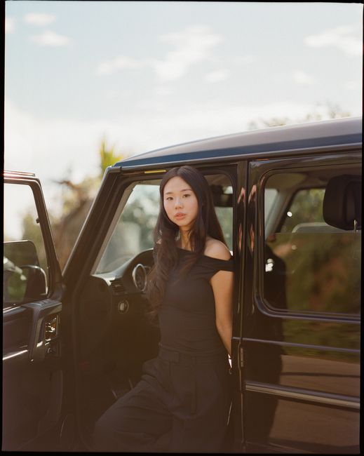 Elegant young woman in black outfit next to a luxury black car outdoors.