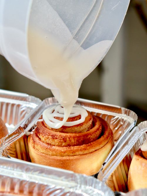 Free A detailed shot showing icing being poured over freshly baked cinnamon rolls in aluminum trays. Stock Photo