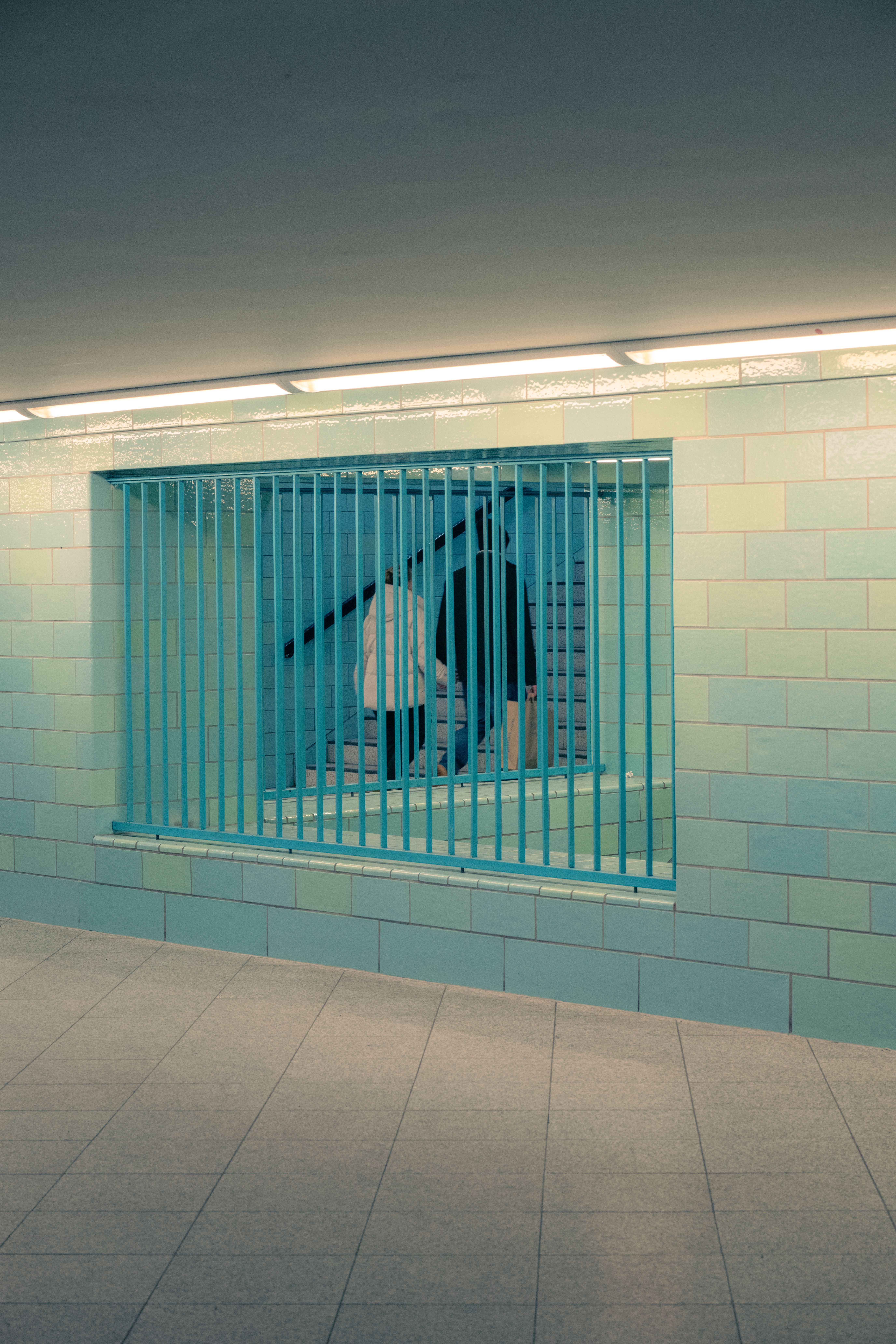 A dimly lit underground hallway featuring a blue barred window with silhouettes on the stairs.