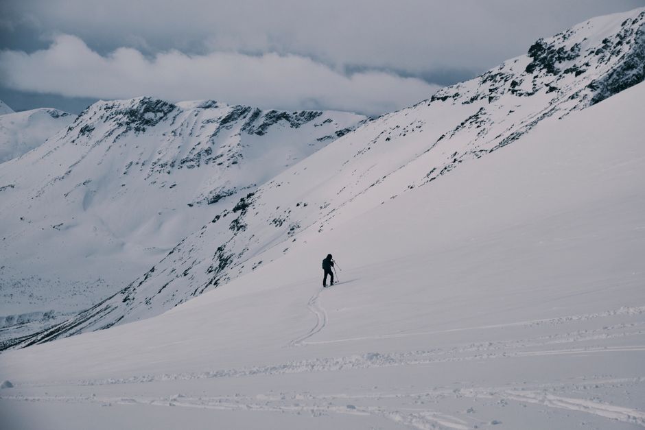A lone skier traversing a vast, snow-covered mountain range under a cloudy sky.