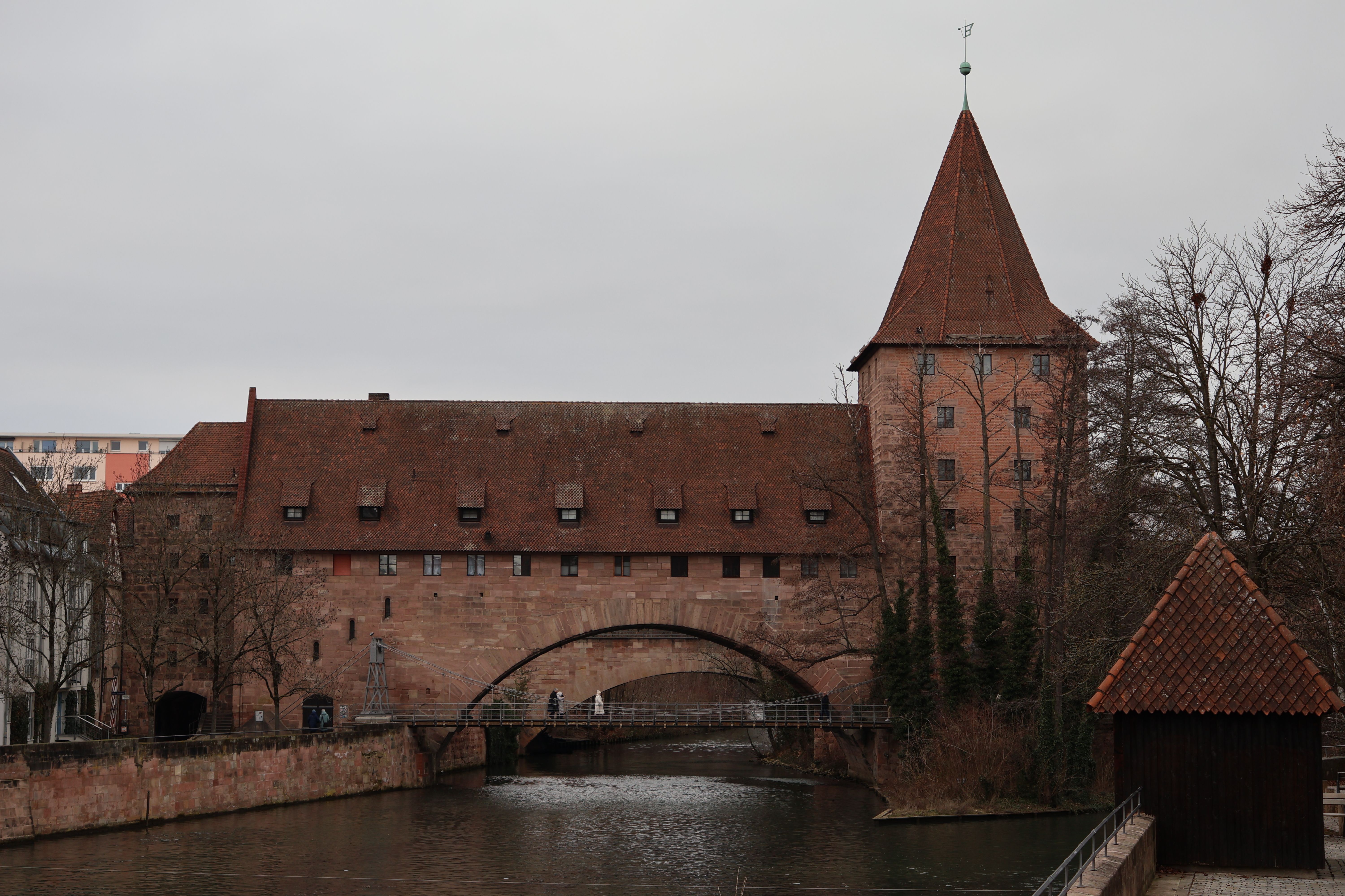 A long red sandstone building with a steep pointed tower, straddling a river with a low arch, under a flat grey sky and bare trees