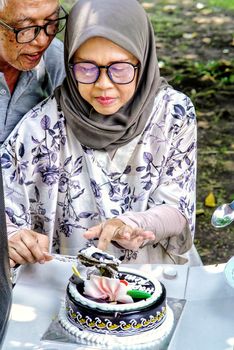 Senior couple cutting a cake outdoors in West Java, Indonesia, enjoying a special occasion.