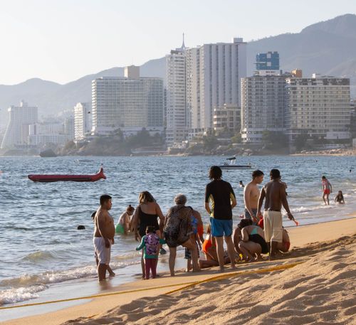 Free Vacationers enjoy the sunny beach in Acapulco with towering city buildings in the background. Stock Photo