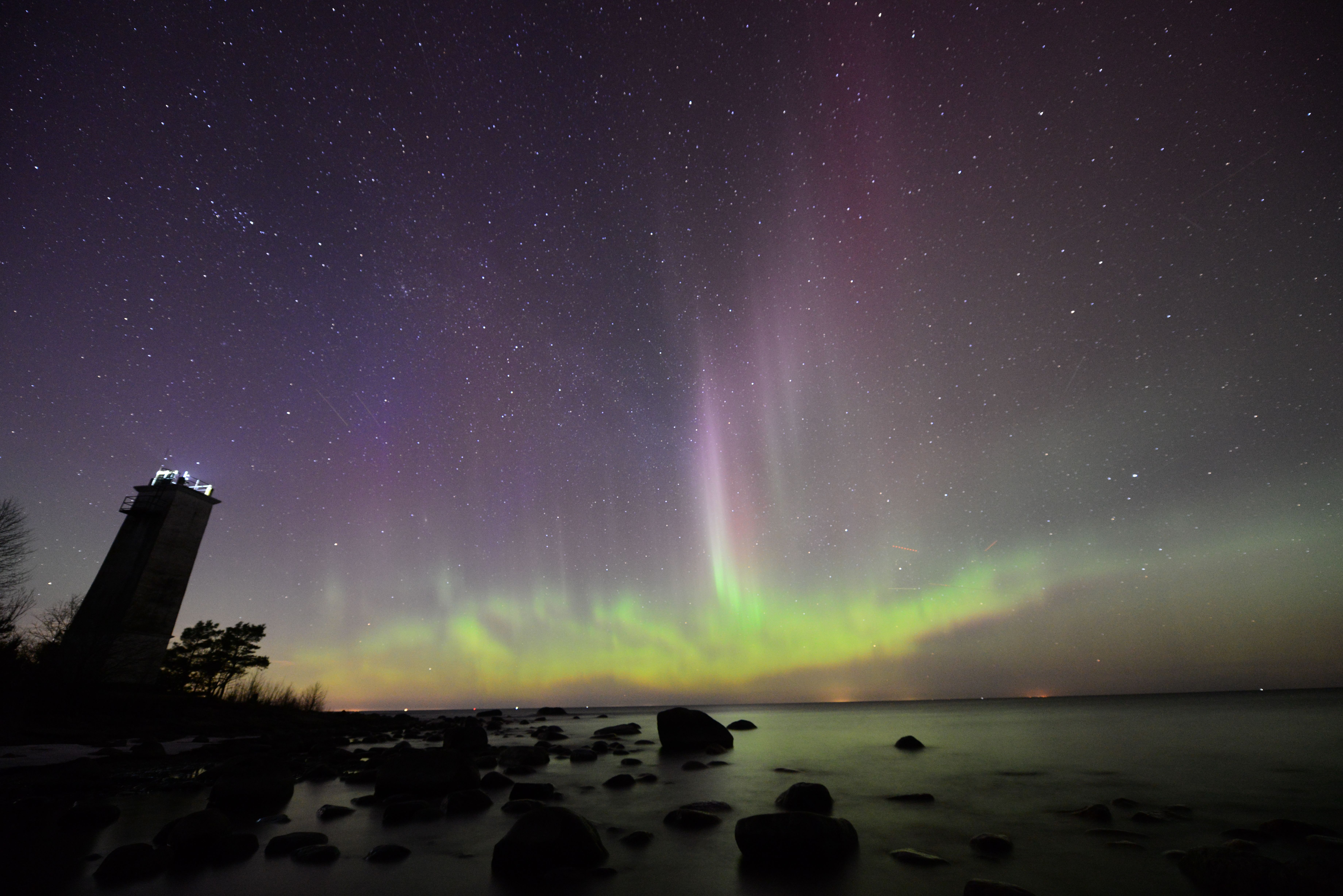 Spectacular view of Northern Lights illuminating the sky over a rocky beach and lighthouse at night.