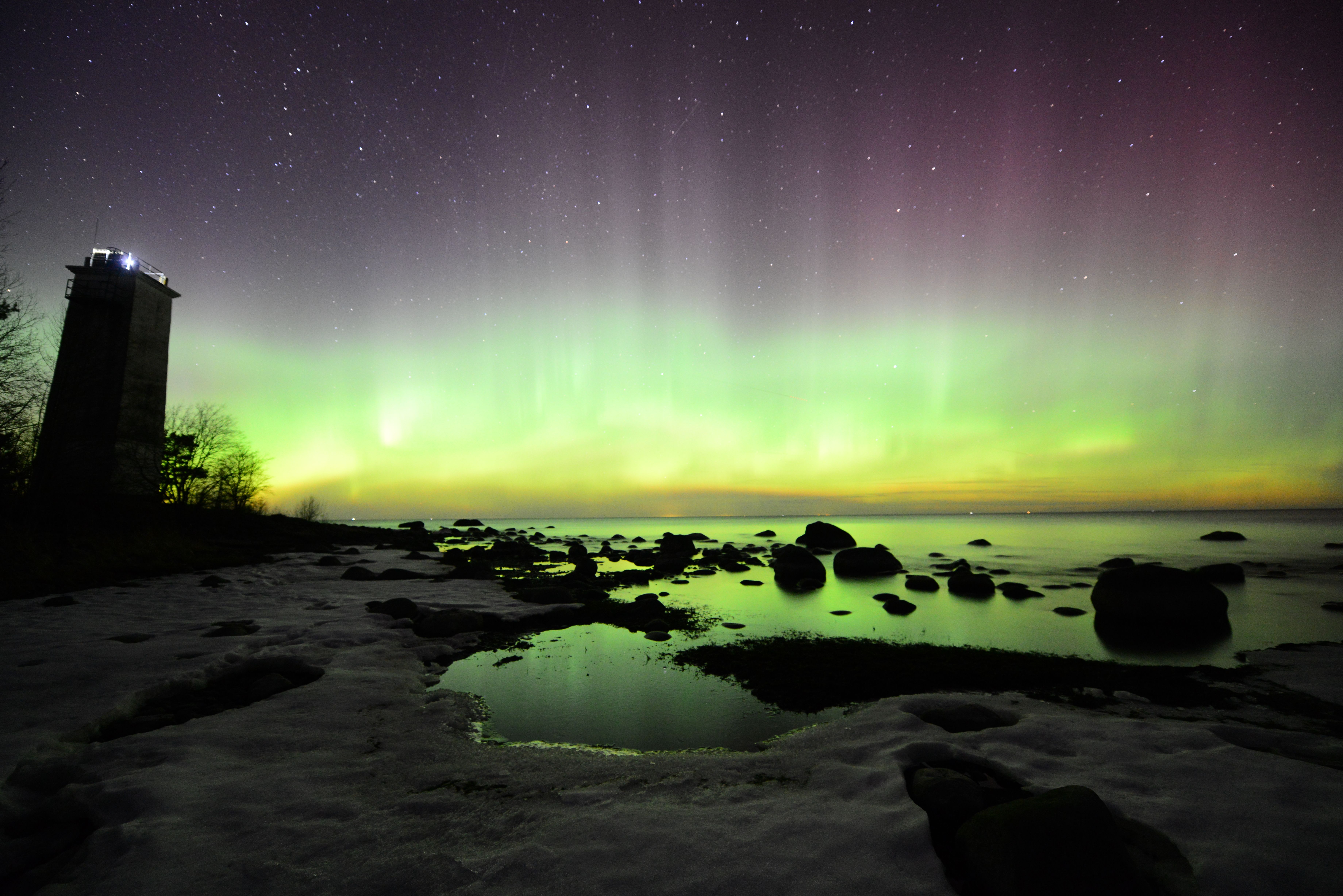 Magnificent aurora borealis illuminates the night sky on a rocky shore with distant lighthouse.