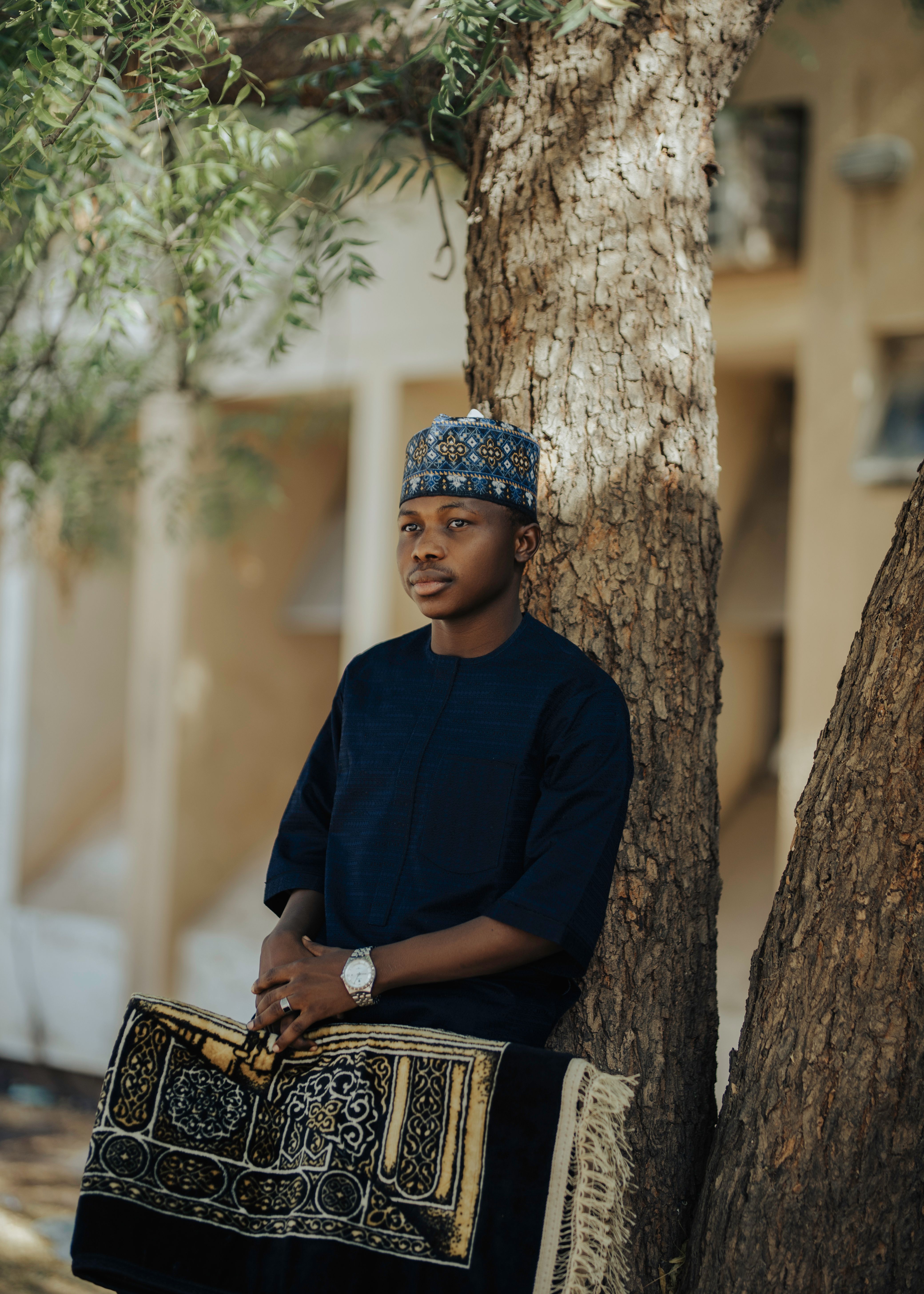 A young man in traditional clothing leaning on a tree outdoors, exuding a calm and reflective mood.