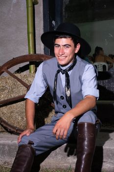 A man wearing traditional gaucho clothing sits by a hay bale, smiling warmly.
