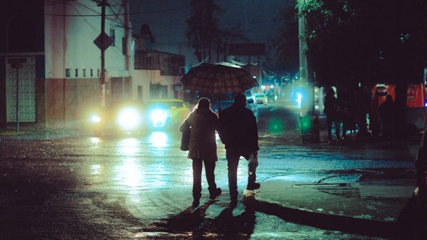 A couple walks under an umbrella on a rainy night in Toluca, Mexico, with city lights illuminating the street.