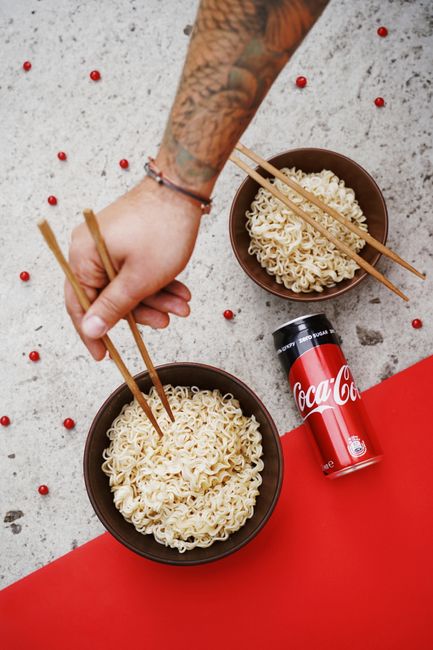 Top view of a tattooed hand using chopsticks to pick instant noodles near a can of Coke.