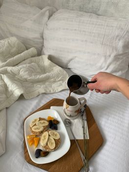 Relaxing breakfast in bed with coffee, fruit, and toast on a wooden tray.
