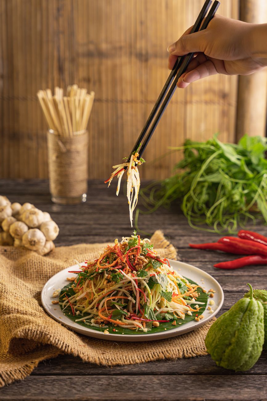 Fresh som tam being prepared in a traditional wooden mortar and pestle, showing green papaya shreds, red chilies, and lime wedges
