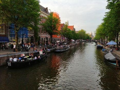 People enjoying a lively evening along Amsterdam's scenic canal with boats and vibrant streets.