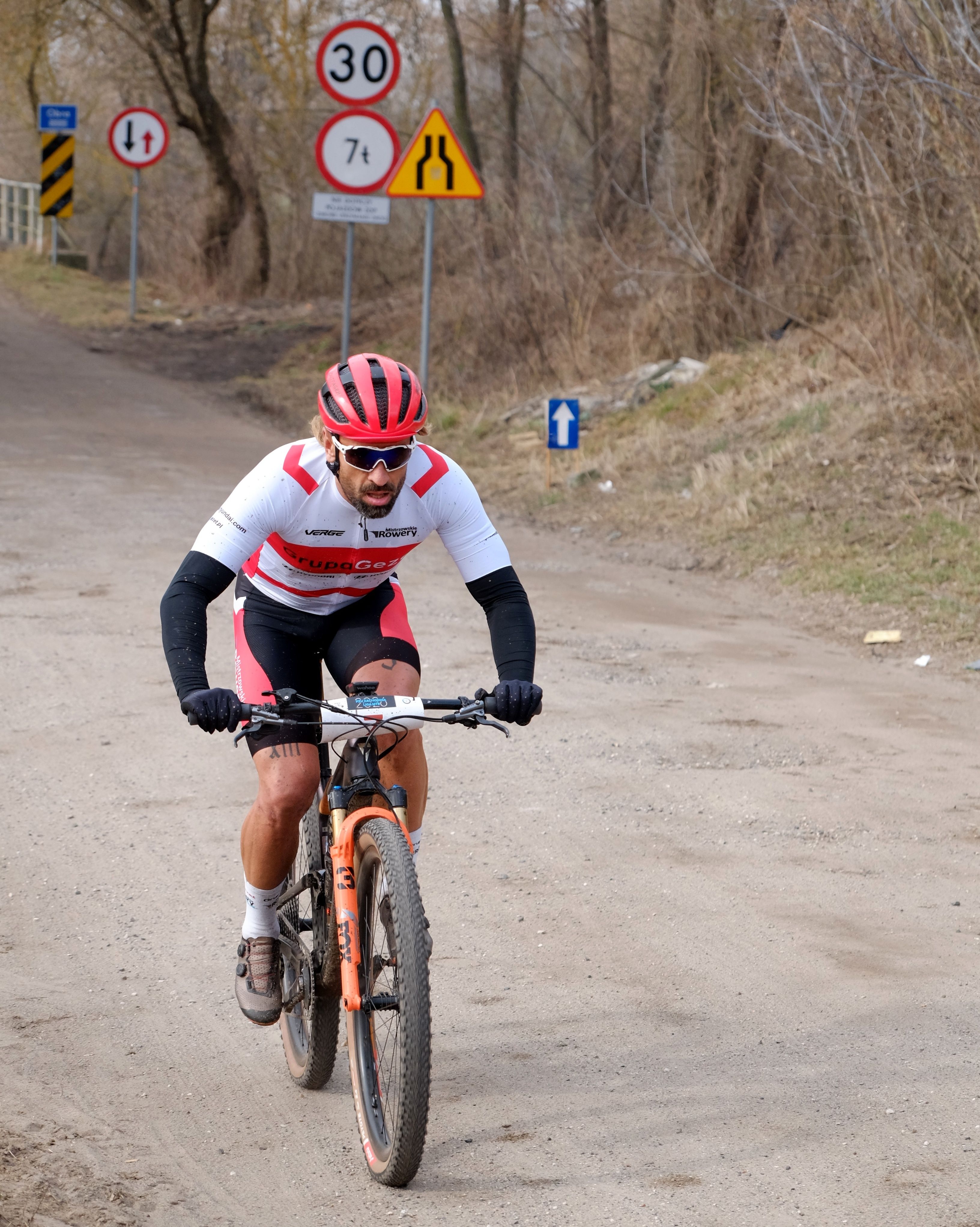 A cyclist wearing red and white sportswear rides on a dirt road near traffic signs, showcasing active lifestyle.