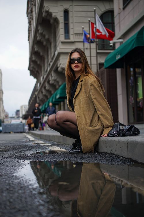 Free Stylish woman in sunglasses and coat posing on street curb with city buildings and flags in the background. Stock Photo