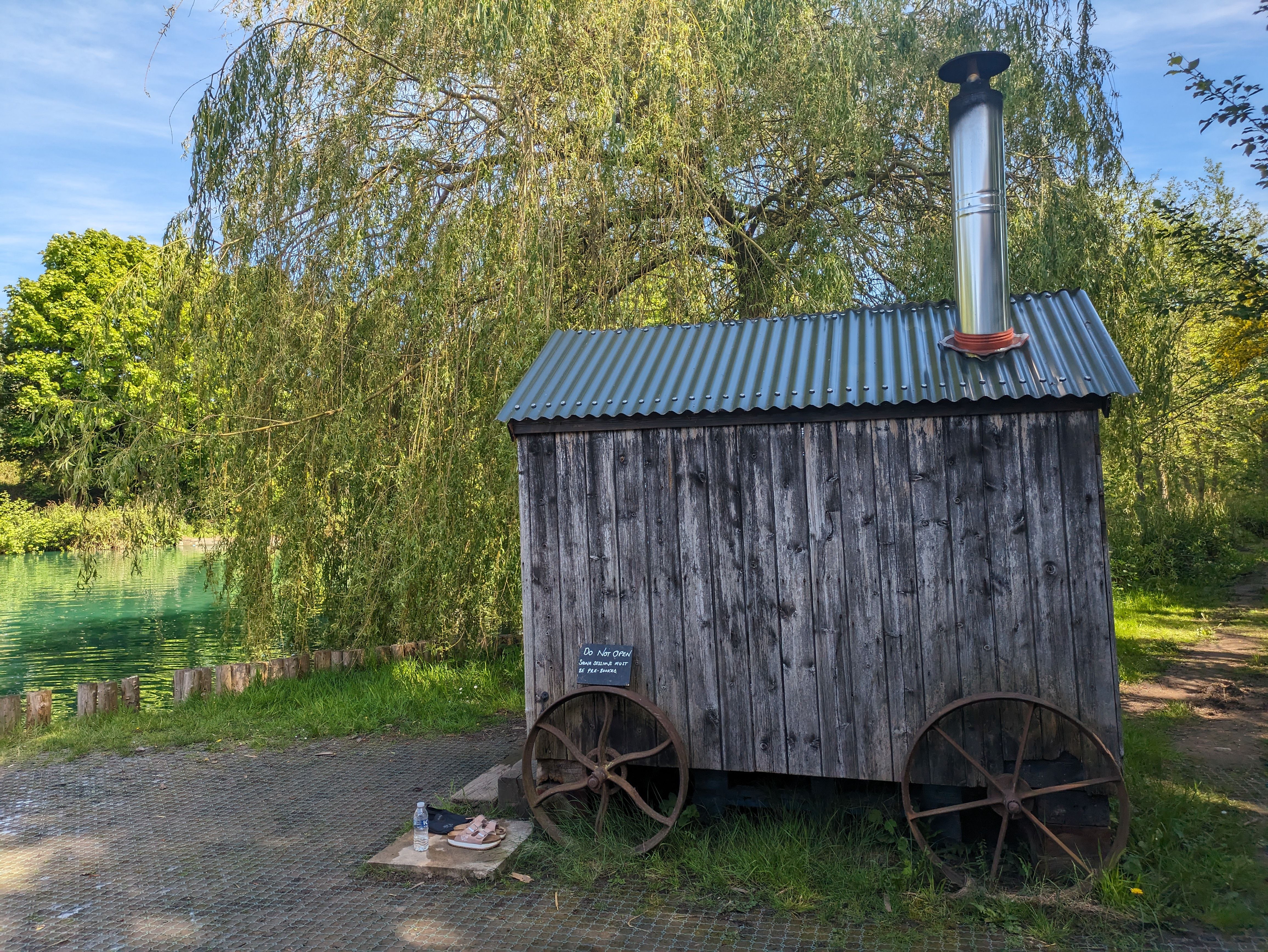 A weathered timber hut with a corrugated roof, a chimney and large iron wheels beside a lake