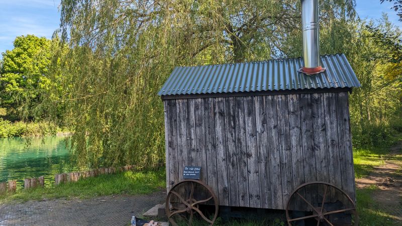 A weathered timber hut with a corrugated roof, a chimney and large iron wheels beside a lake