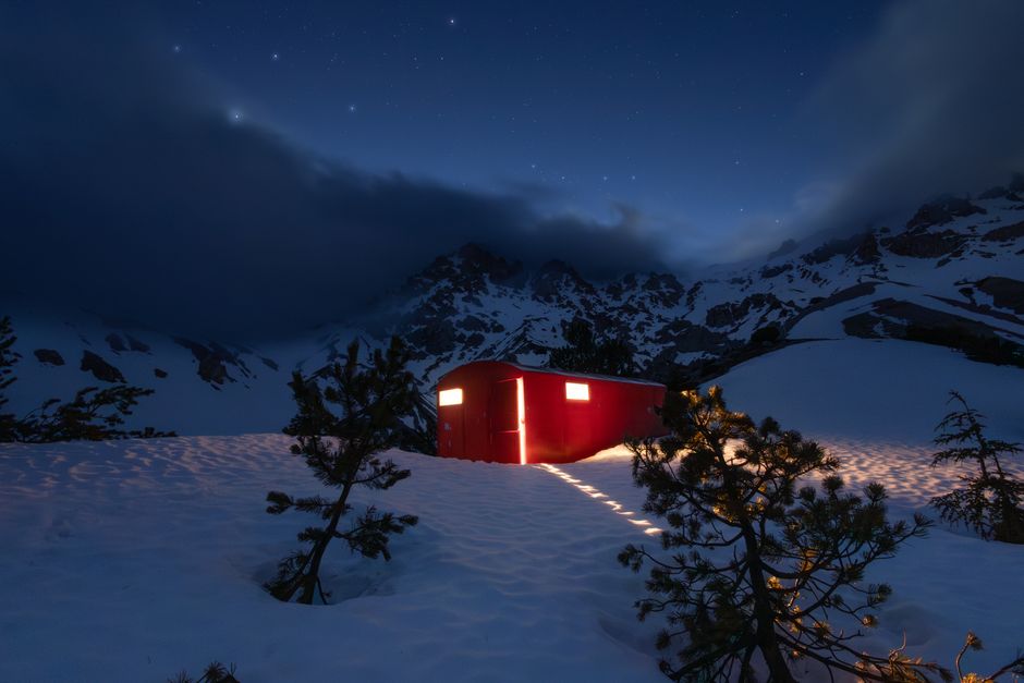 Red mountain hut illuminated under a starry night sky, surrounded by snow and trees.