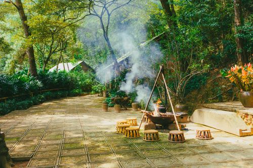 Free Rustic cooking area with a fire pit in a lush forest, featuring stools and smoke rising. Stock Photo