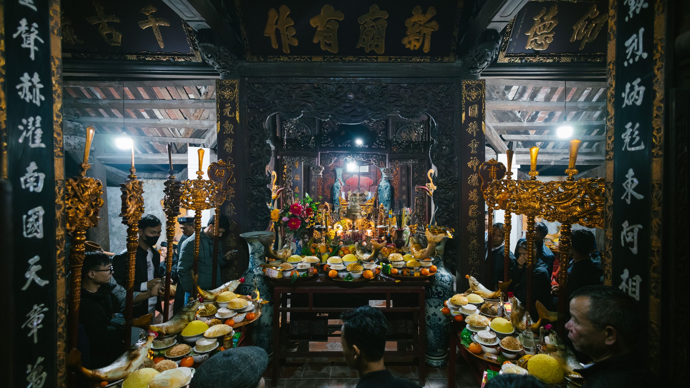 Ceremony inside Asian temple with offerings like fruits and incense, surrounded by traditionally dressed people.