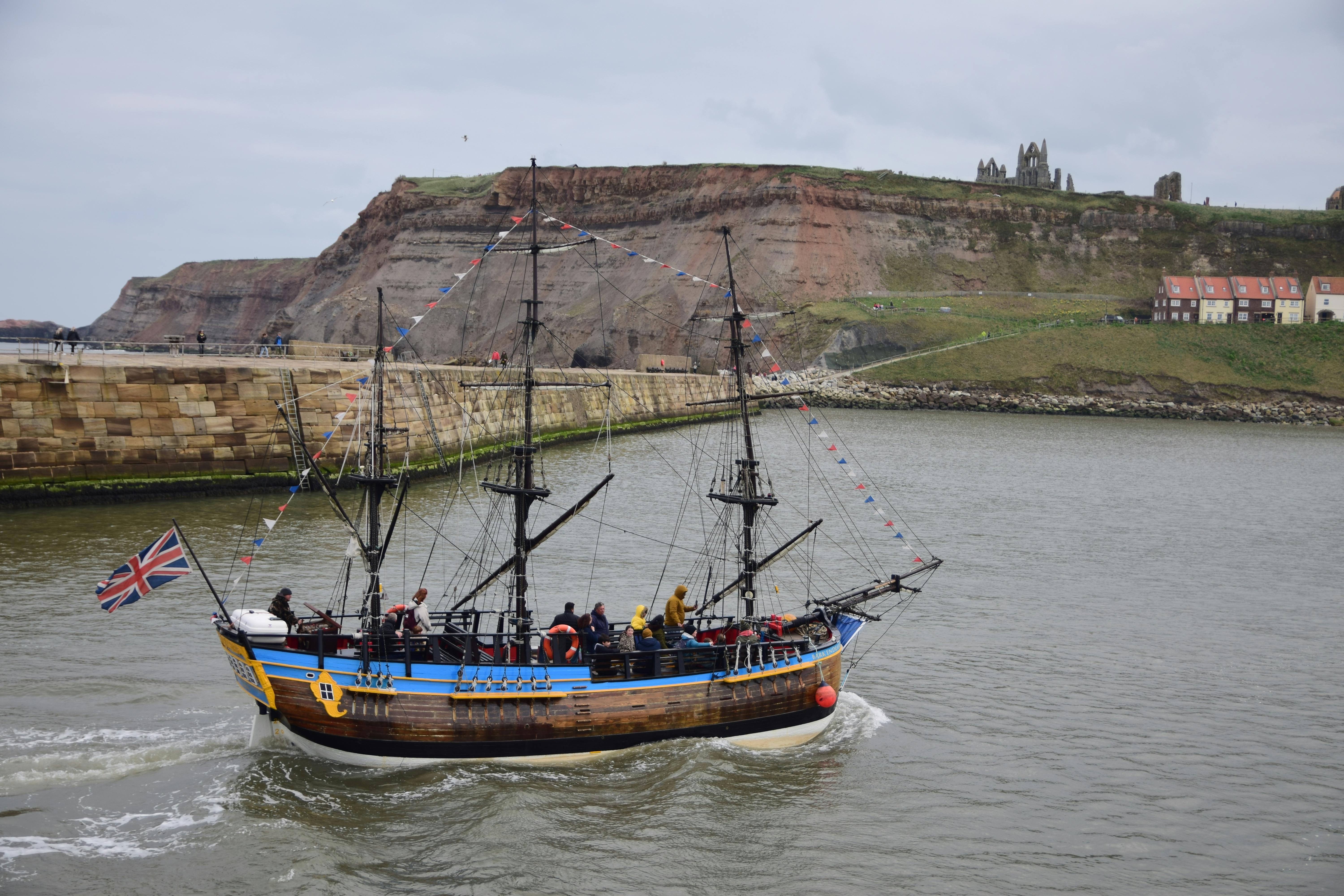 A historic ship sails past Whitby Abbey ruins under overcast skies, showcasing maritime charm.