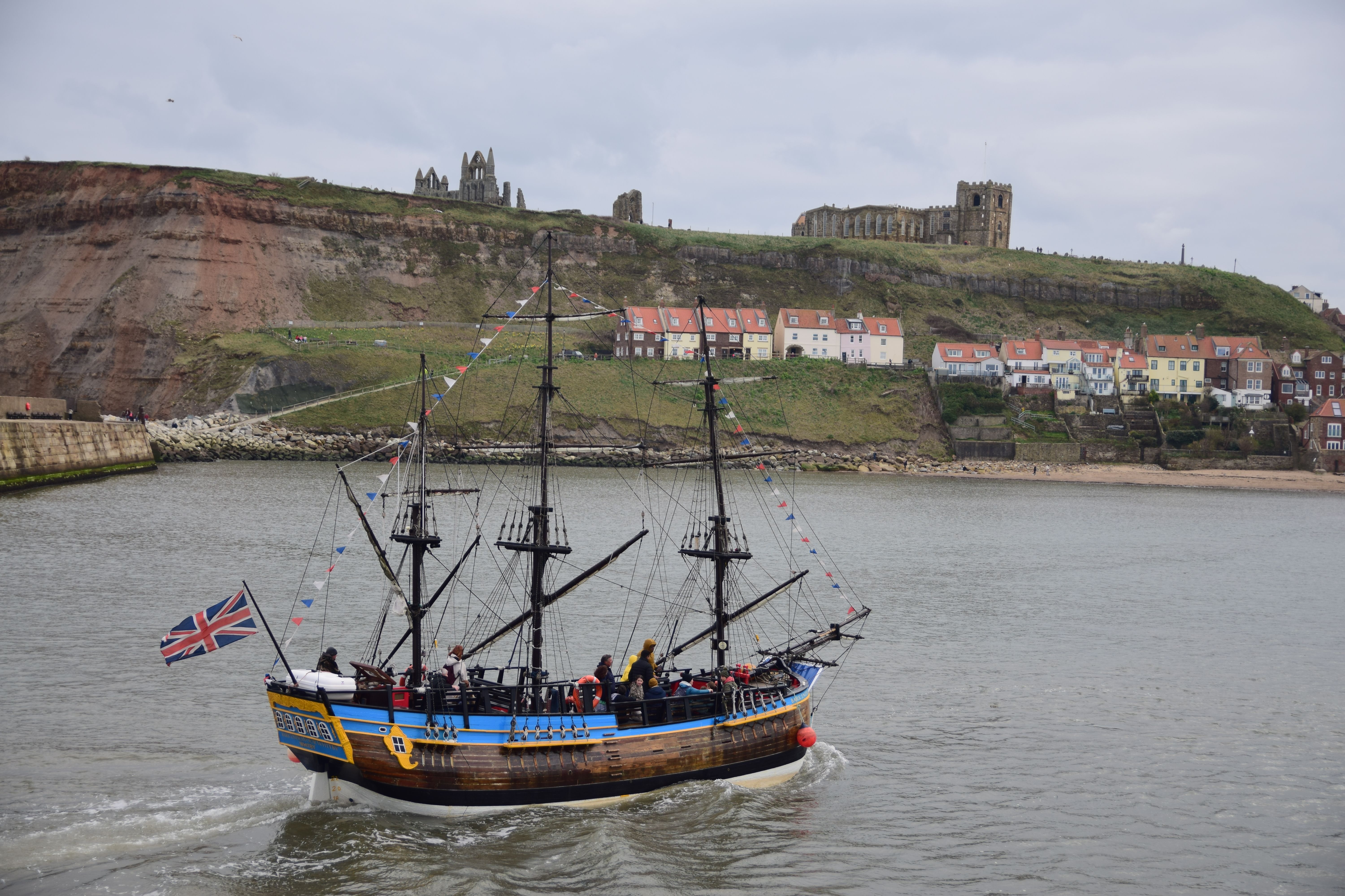 A traditional sailing ship with a British flag navigates near the iconic ruins of Whitby Abbey.