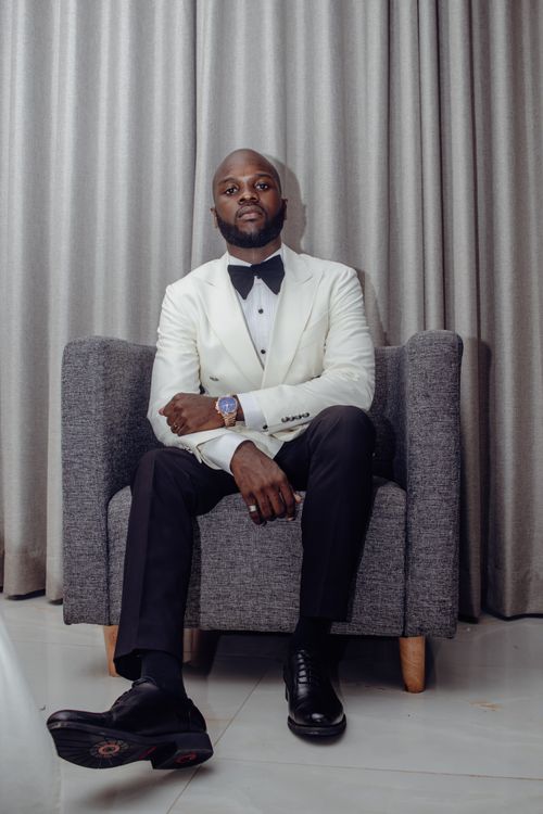 Free Stylish man in a white tuxedo sitting confidently on a gray chair indoors. Stock Photo