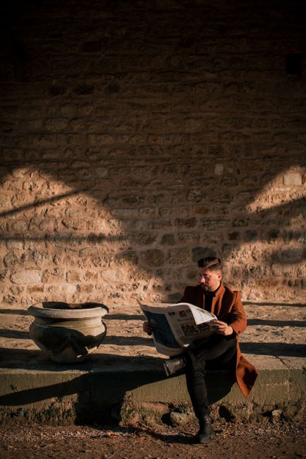 Stylish man reading a newspaper on a stone bench in sunlight.