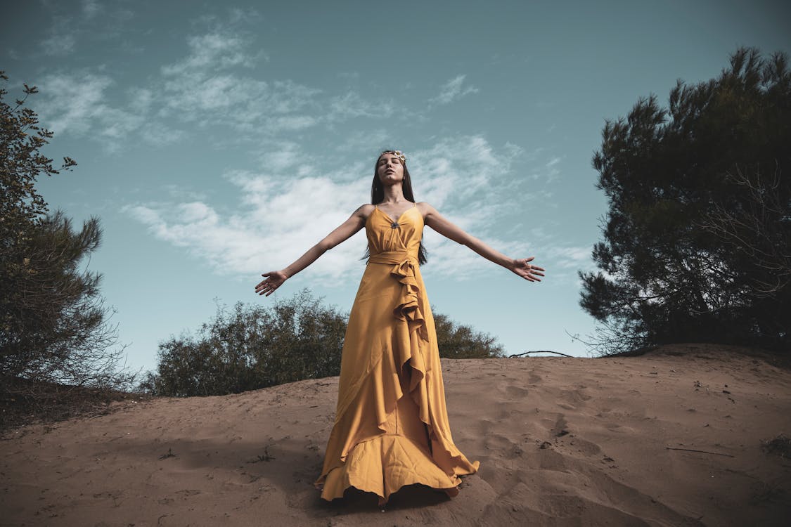 Free Photo of Woman in Yellow Dress Standing on Brown Sand Under Blue Sky with her Eyes Closed and Her Arms Spread Stock Photo