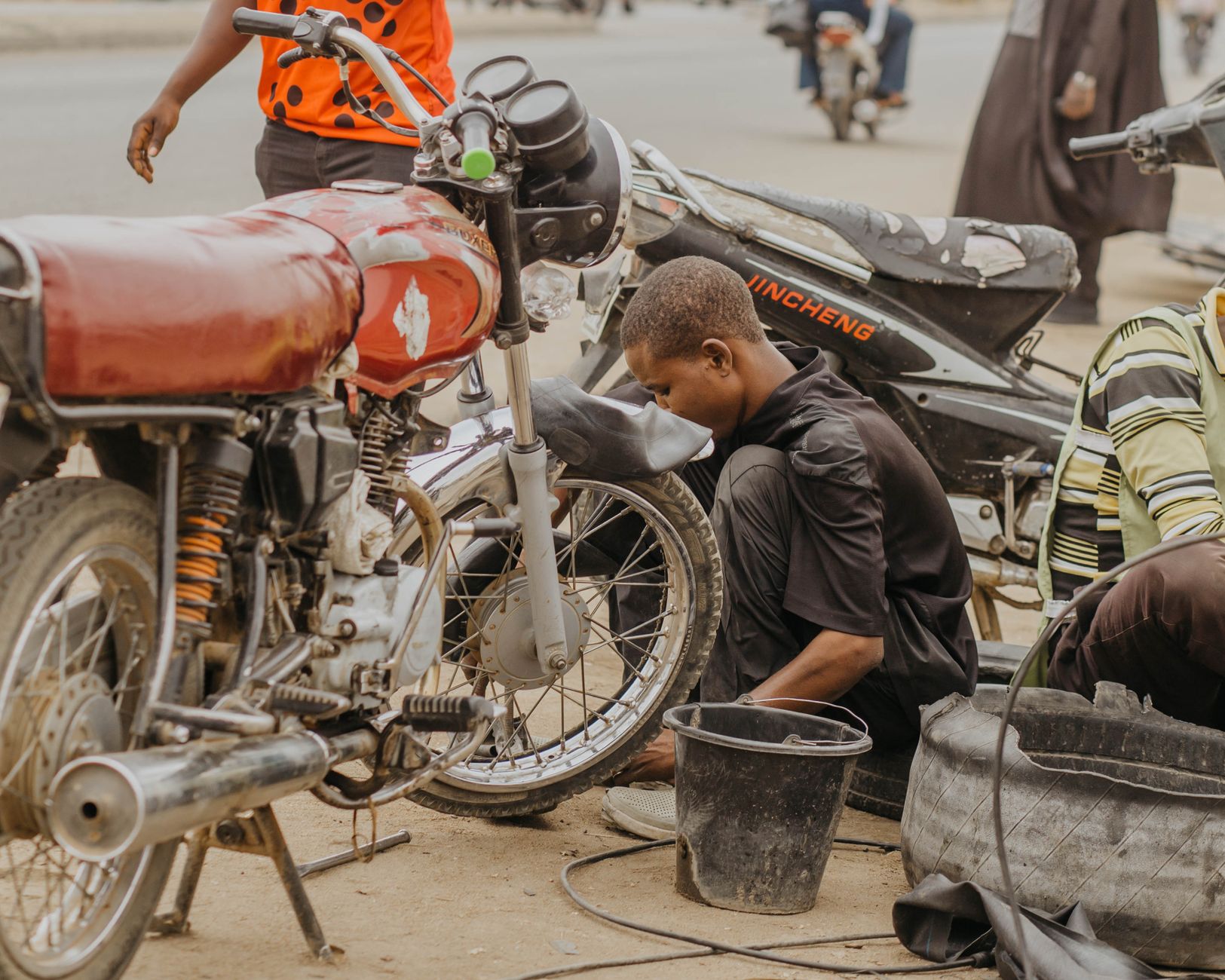 A young mechanic repairs a motorcycle on a busy street, showcasing urban life and local industry.