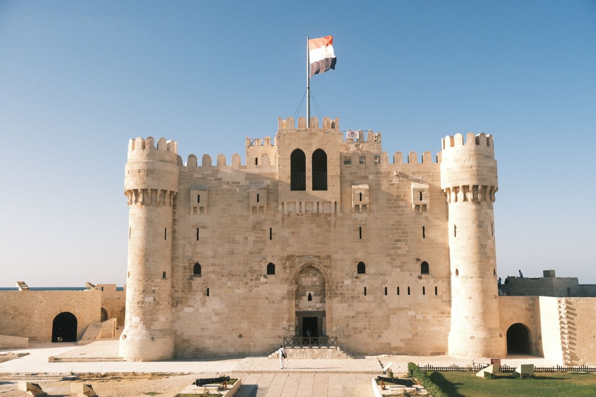The Citadel of Qaitbay head-on, a pale limestone fortress with twin round towers and an Egyptian flag flying against a clear blue sky.