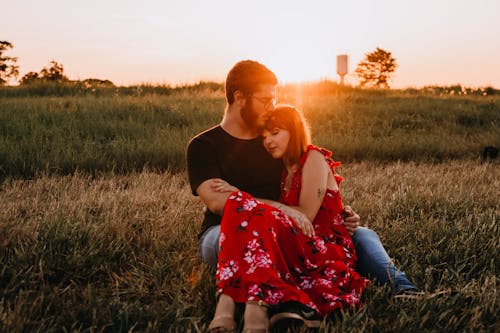 Man In Zwarte Ronde Hals T Shirt Zittend Op Grasveld Naast Vrouw In Rode Bloemen Jurk