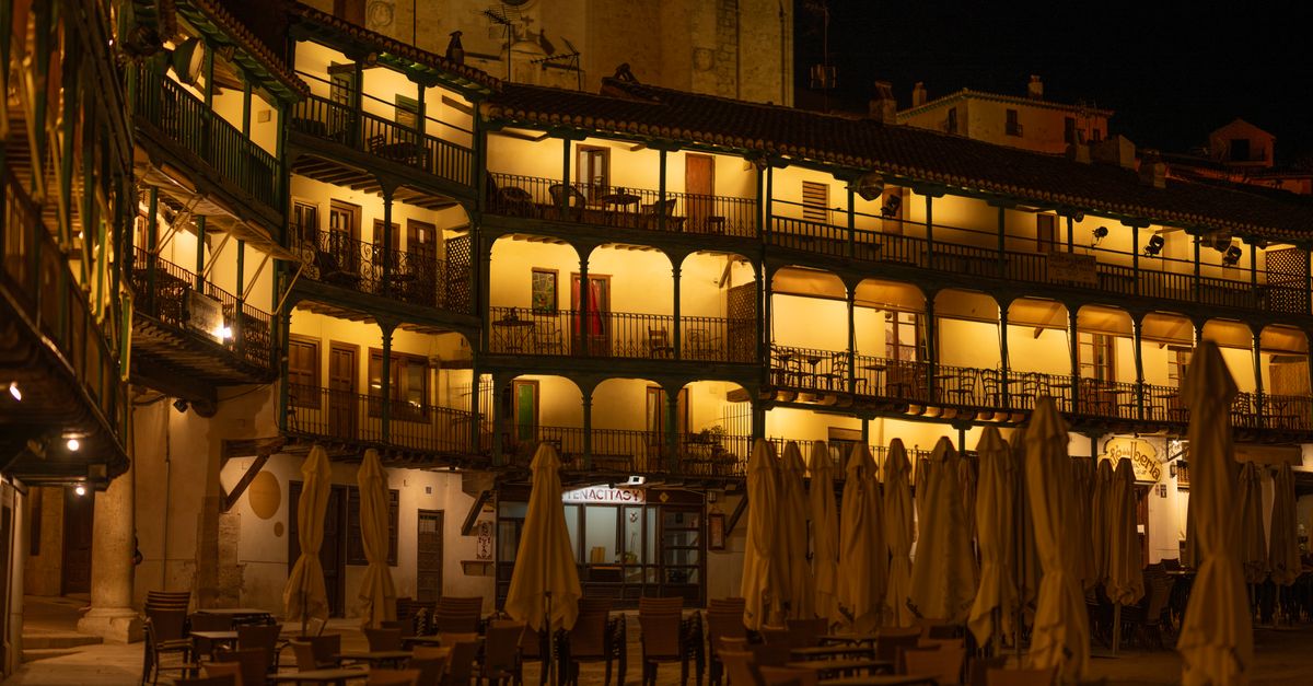 Illuminated arched balconies in Chinchón's Plaza Mayor with closed umbrellas and empty chairs creating a moody night scene.