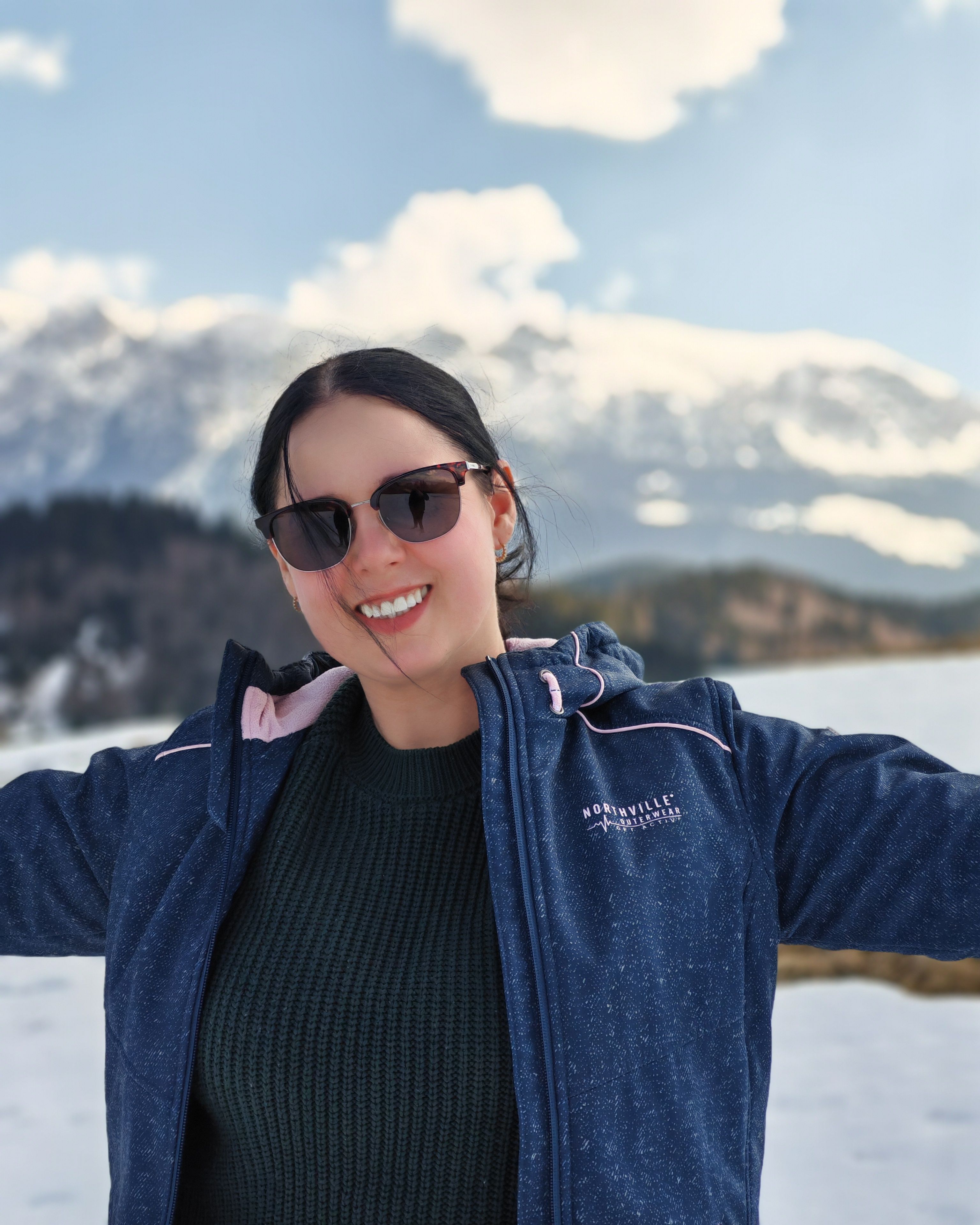 A smiling woman in sunglasses enjoys a winter day with snowy mountains in the background.