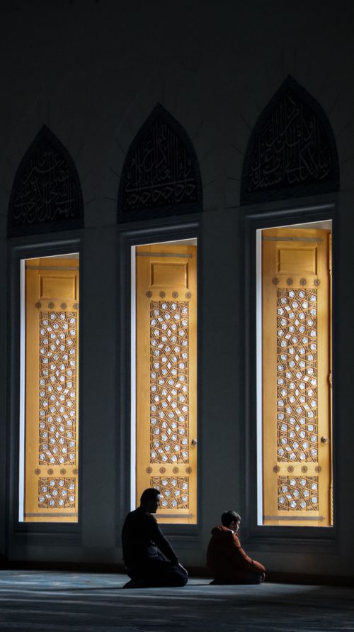 Free Silhouettes of a man and child praying by illuminated ornate mosque windows at night. Stock Photo