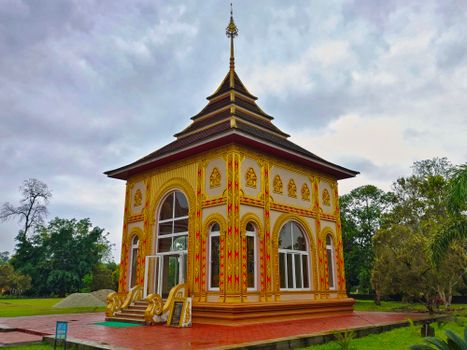 An intricately designed pavilion with golden details in a tropical garden setting, under overcast skies.