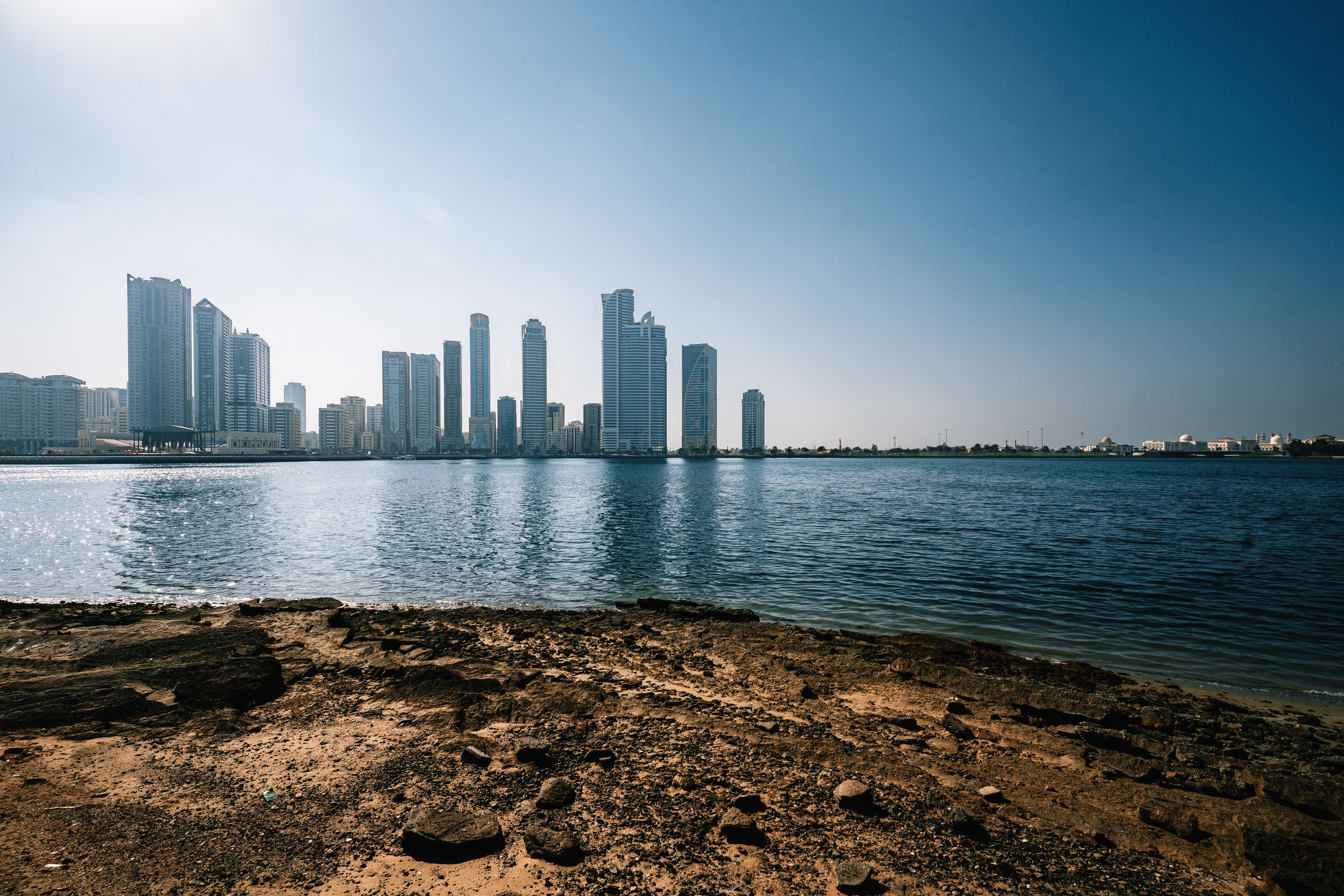 Scenic view of Sharjah skyline reflecting on the calm water under a clear blue sky.