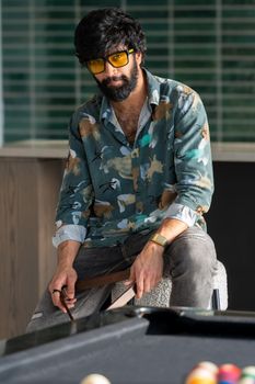Man with beard and sunglasses playing pool in a casual setting.