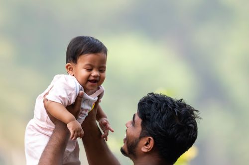 Free A father joyfully lifts his smiling baby in an outdoor setting, capturing a moment of pure happiness. Stock Photo