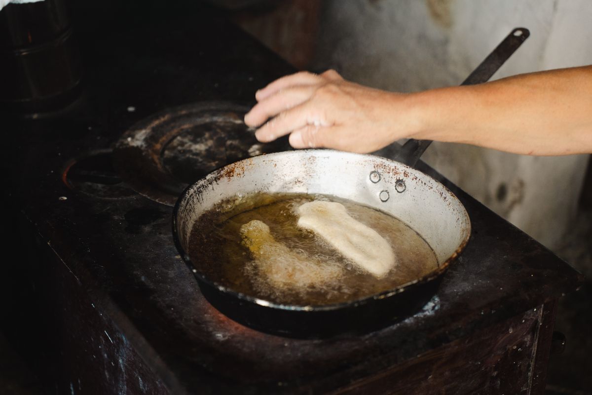 A hand over a pan of hot oil on a blackened stove as a piece of dough fries in a home kitchen