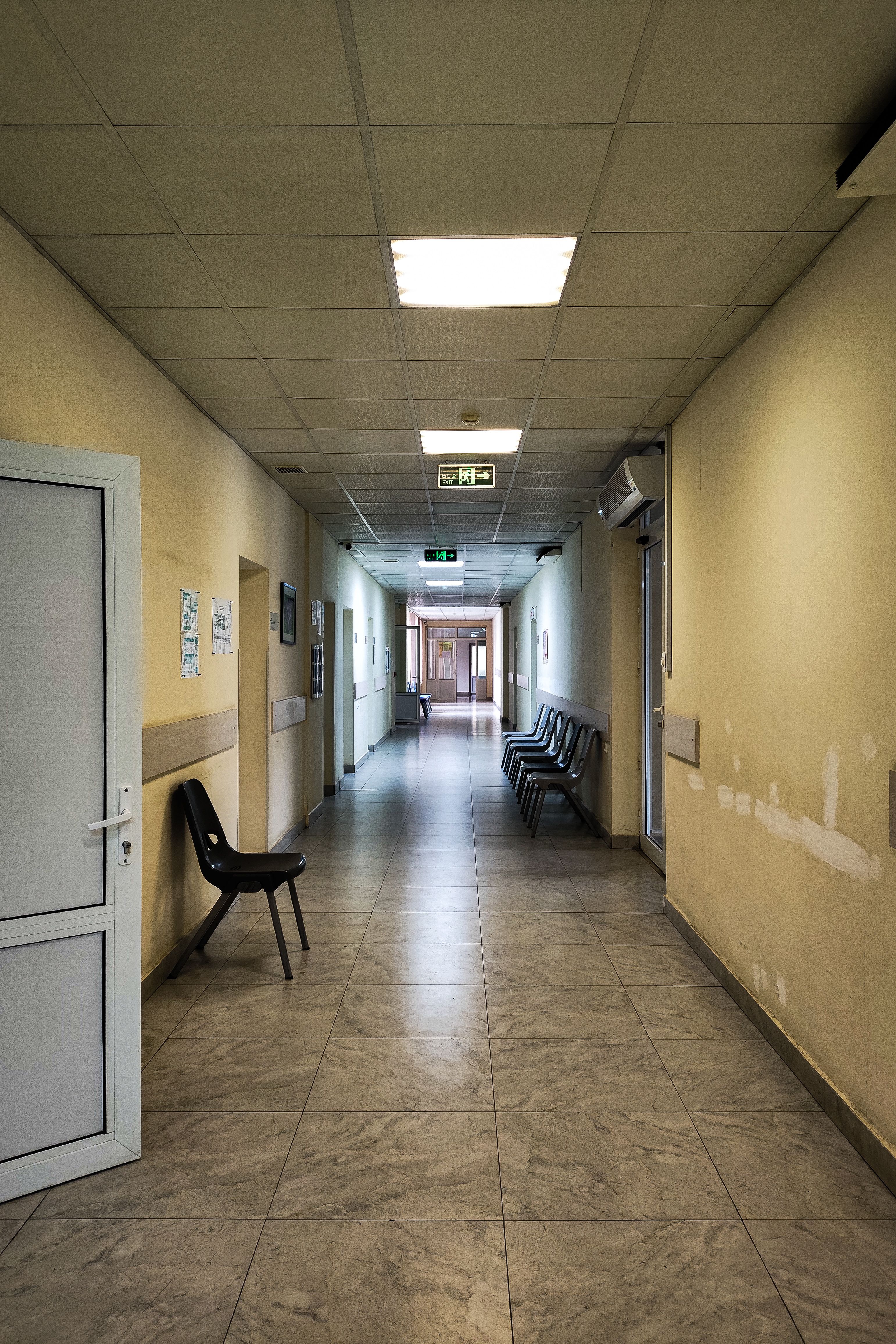 An empty hospital corridor with open doors and chairs, lit by fluorescent lights.