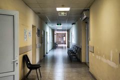 An empty hospital corridor with open doors and chairs, lit by fluorescent lights.